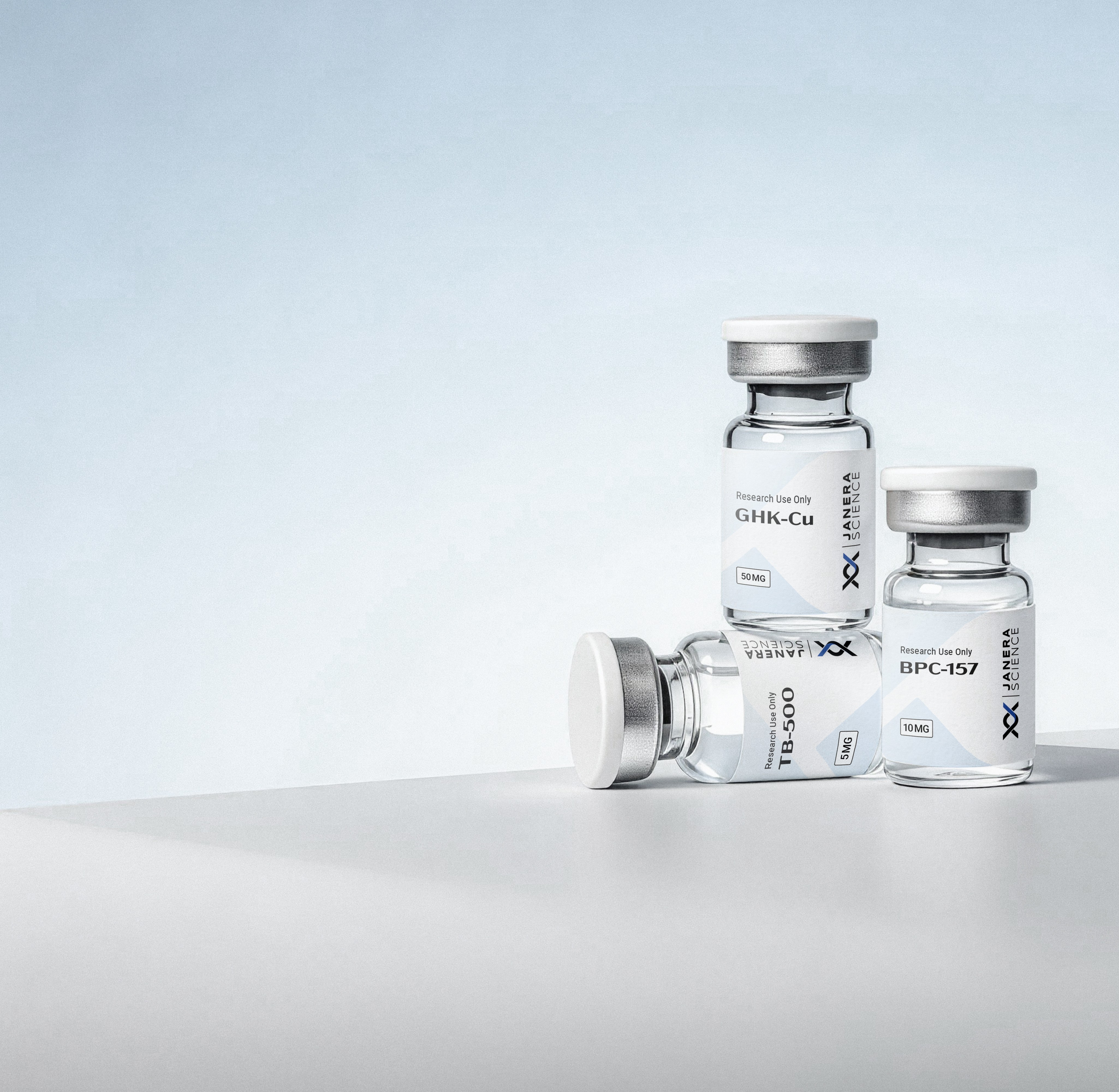 Three Janera Science research peptide vials — GHK-Cu 50mg standing upright at center rear, BPC-157 10mg to the right, and TB-500 5mg lying flat in the foreground — arranged on a light gray surface against a pale blue-gray background.