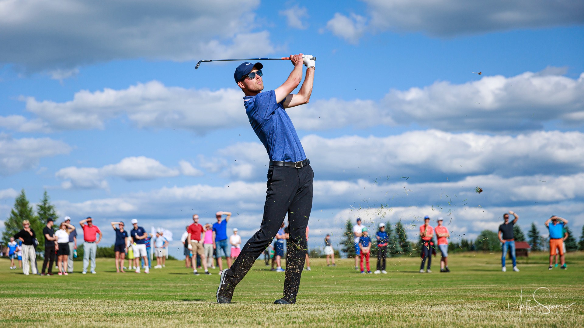 Mark Suursalu mid-swing at Niitvälja Golf, grass debris flying, gallery of spectators behind. Photo by Mats Soomre, 2019.
