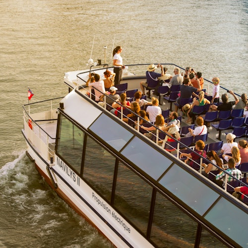 A boat named "NAVILYS II" with many passengers seated, cruising on a river under clear weather.
