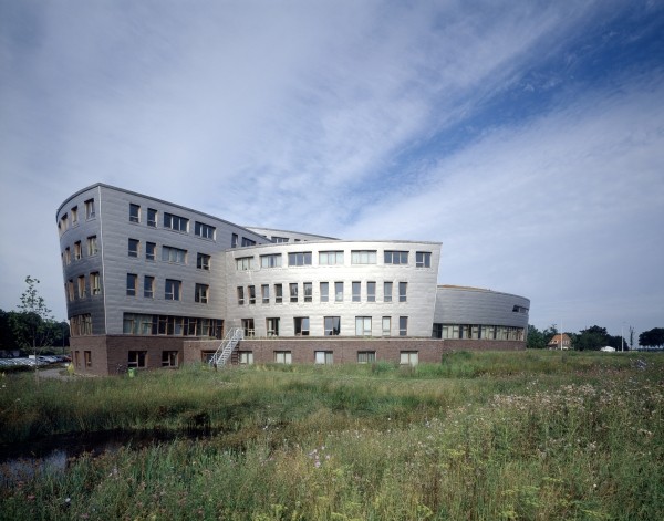 A modern, curved office building with a mix of brick and metal exterior stands amidst lush green grass and wildflowers under a partly cloudy sky.