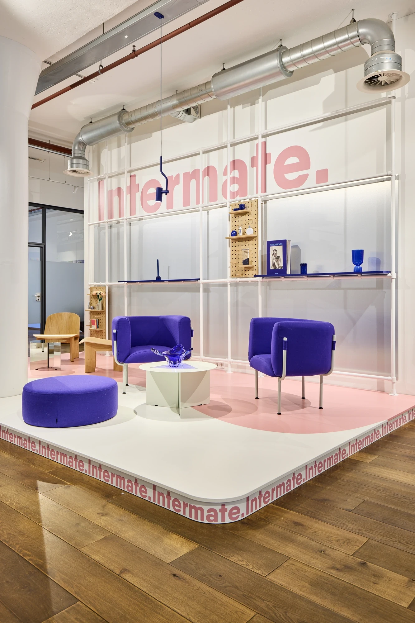 Intermate Hamburg office lounge stage with pink floor inlay, „Intermate." logo on the wall, three purple armchairs and a styled product shelf.