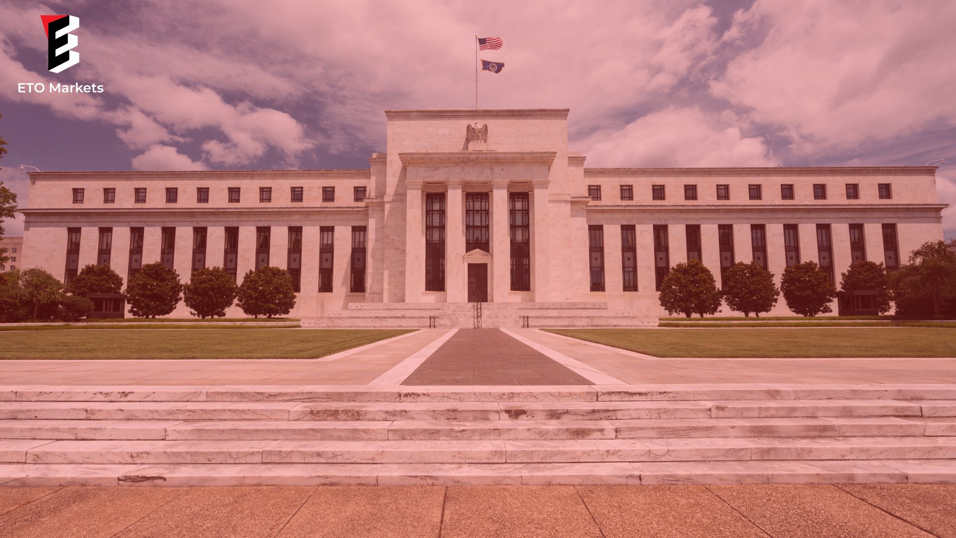 The US Federal Reserve Building with a flag on top, surrounded by green trees and a wide stone walkway leading to its entrance.