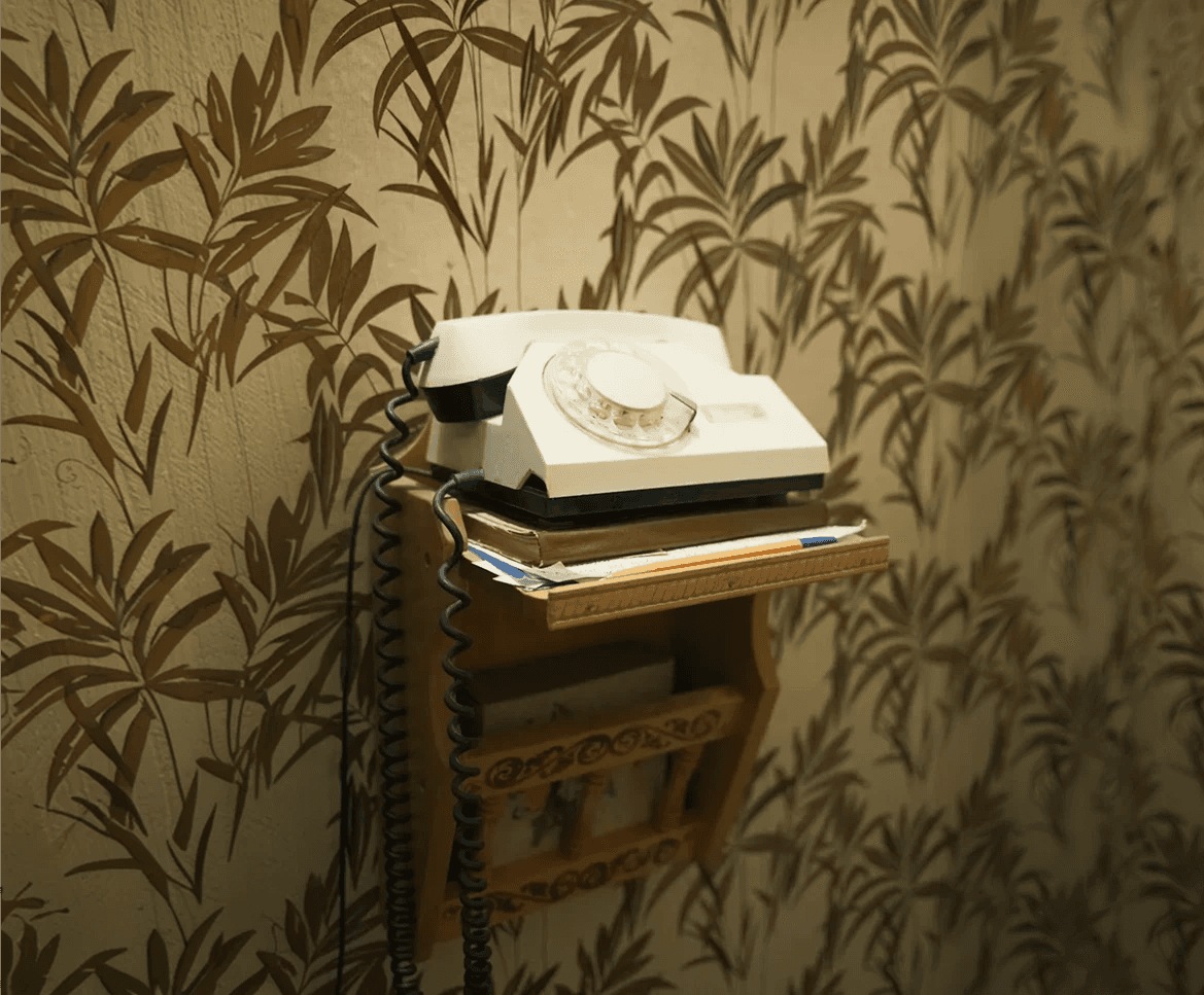 Vintage rotary telephone placed on a small wooden shelf, with papers stacked underneath and patterned wallpaper in the background.