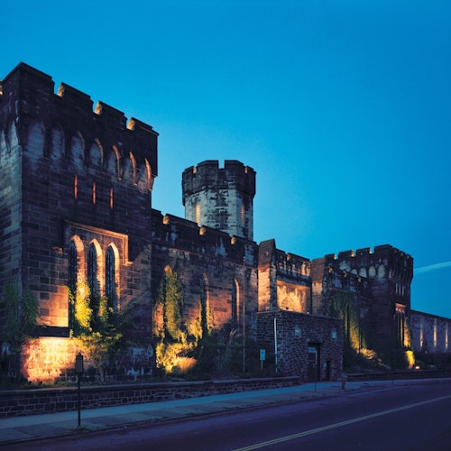 A large, illuminated stone castle with multiple turrets and towers at dusk, viewed from a nearby street.
