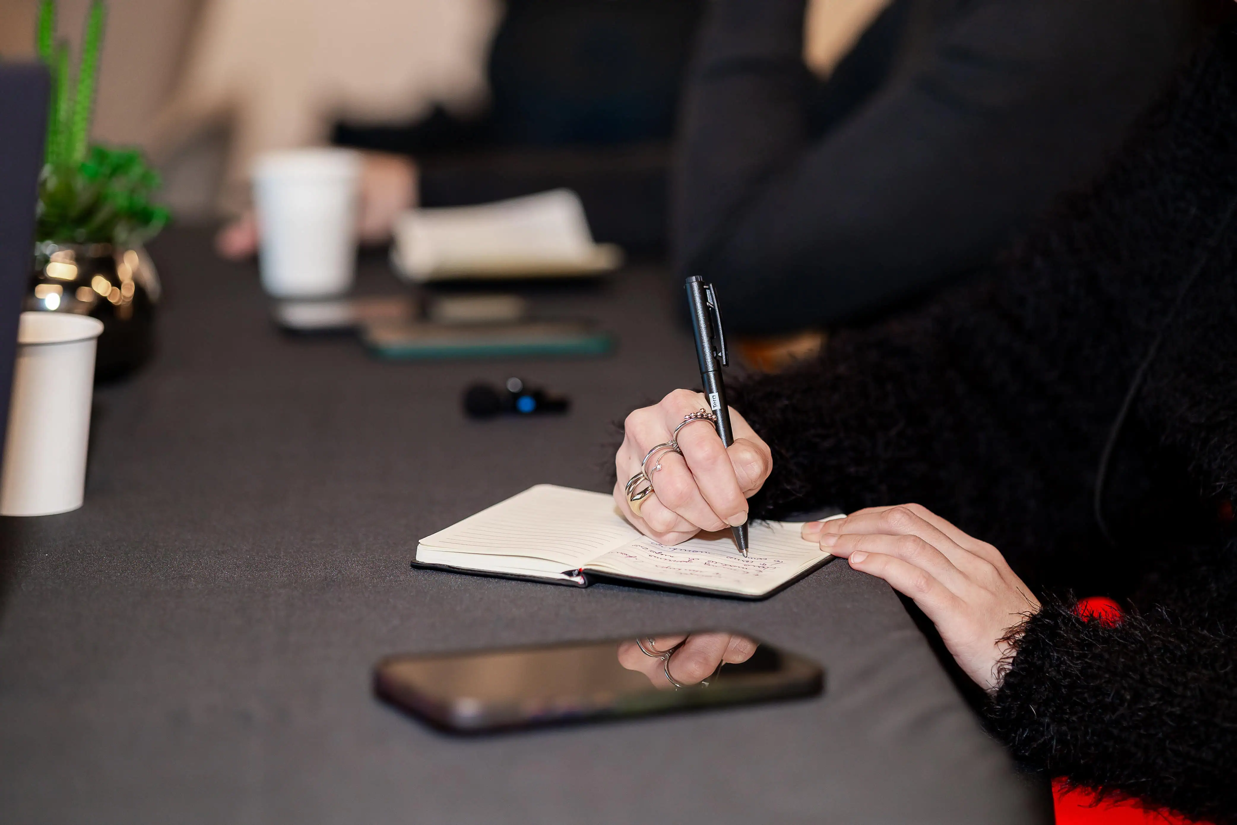 Uma pessoa escrevendo em um caderno sobre uma mesa com uma xícara e outros objetos em um ambiente profissional.