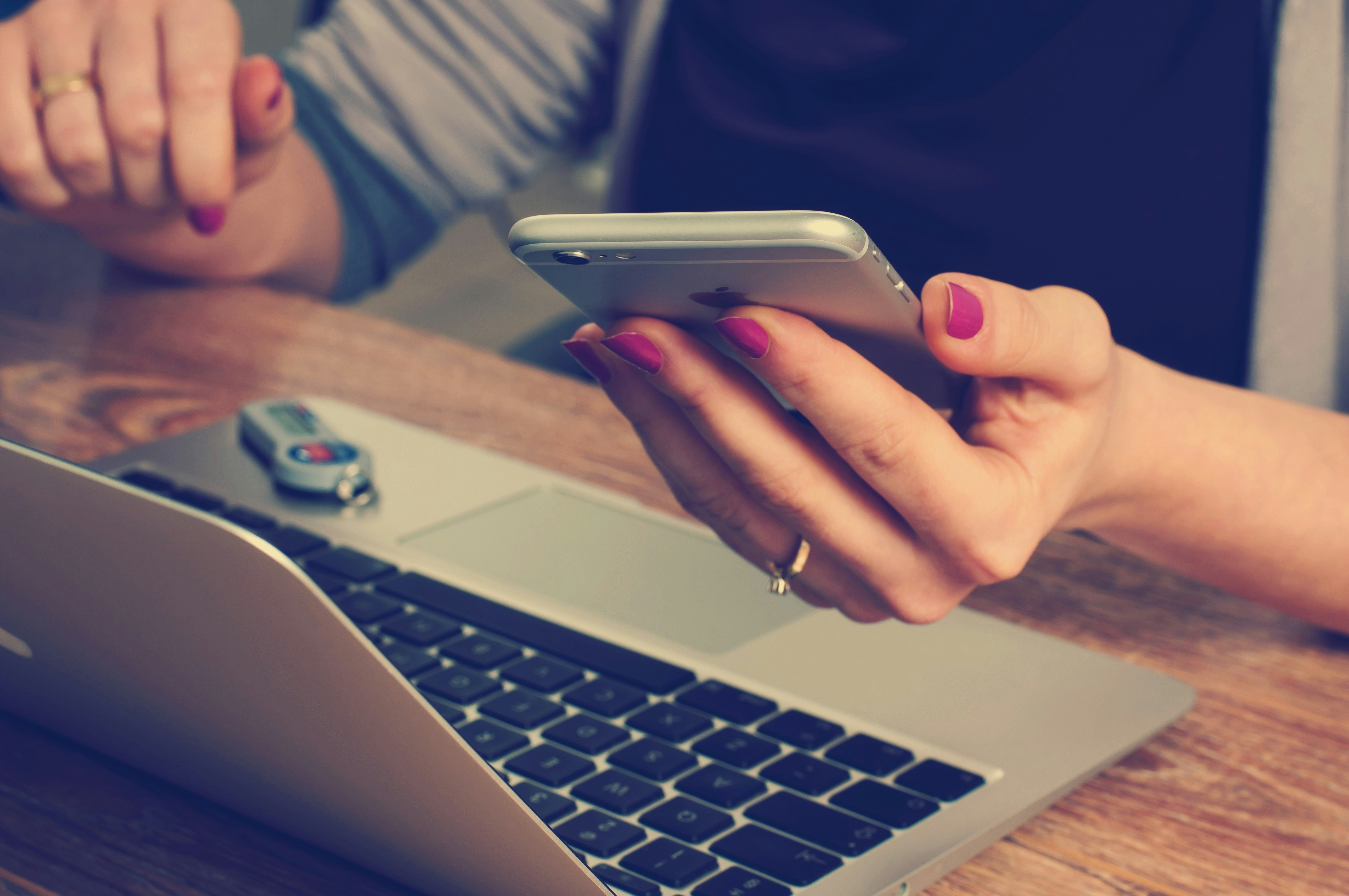 A lady holding a mobile phone in front of her laptop at a wood desk. 