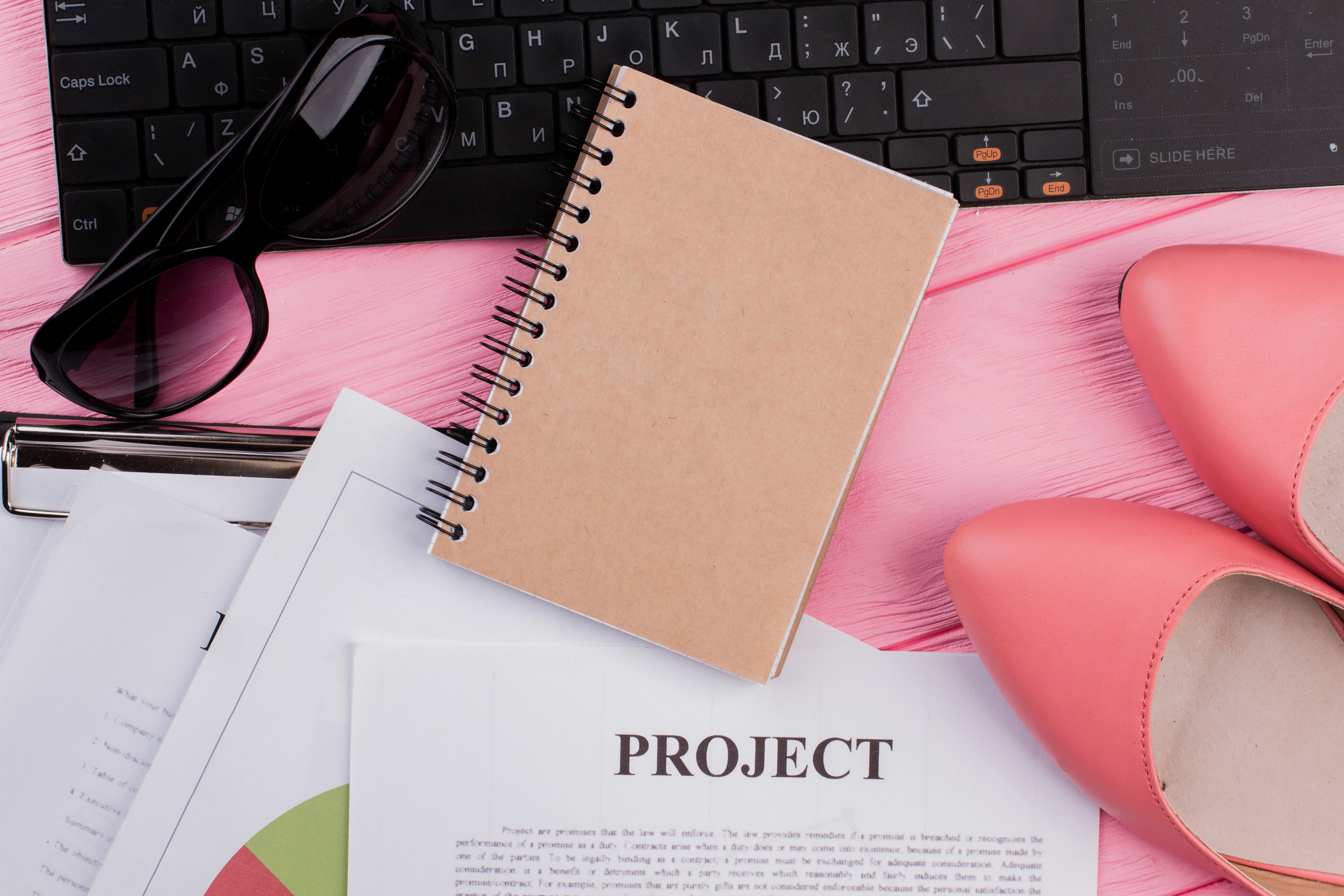 “Flat lay of a spiral notepad with fashion accessories, black sunglasses, a computer keyboard, and pink high heels on a pink wooden desk — representing creative project planning for fashion or lifestyle brands.”