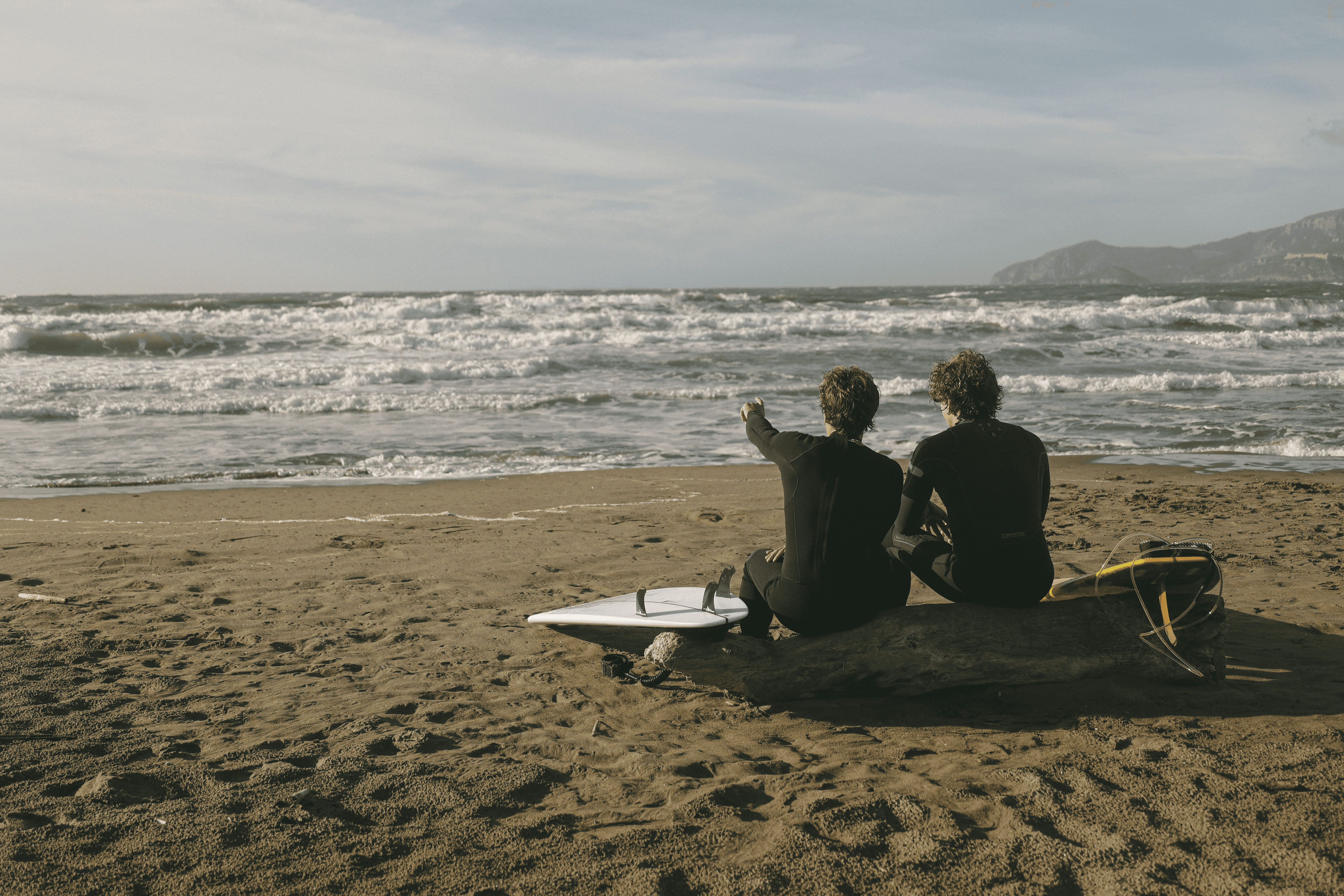 Due surfisti in muta siedono sulla spiaggia, uno indica l'oceano mosso. Due tavole da surf sono appoggiate sulla sabbia.