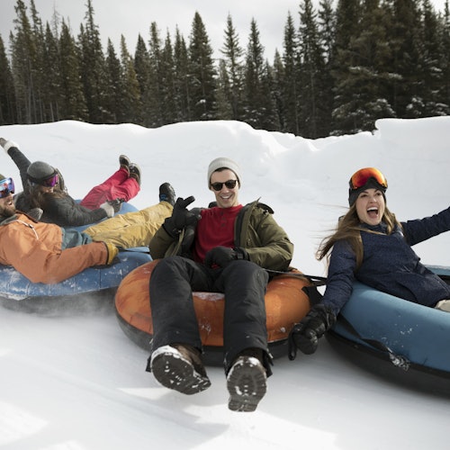 Four people laughing and posing on snow tubes in a wintry forest setting, wearing winter gear and surrounded by snow.