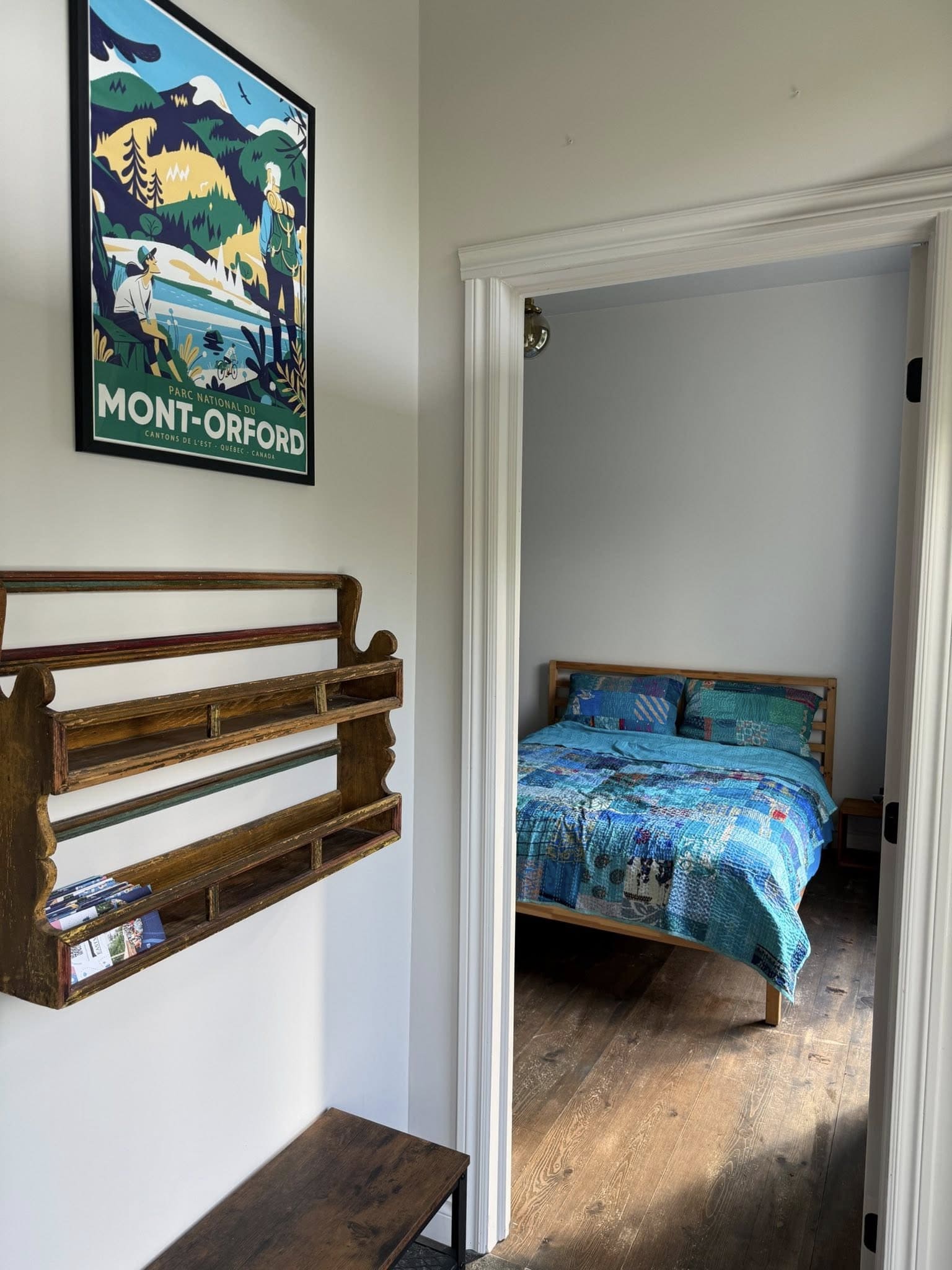 Chambre du gîte R112-Nord avec lit double recouvert d’une courtepointe patchwork bleue et verte, étagère murale en bois et affiche vintage du parc national du Mont-Orford.