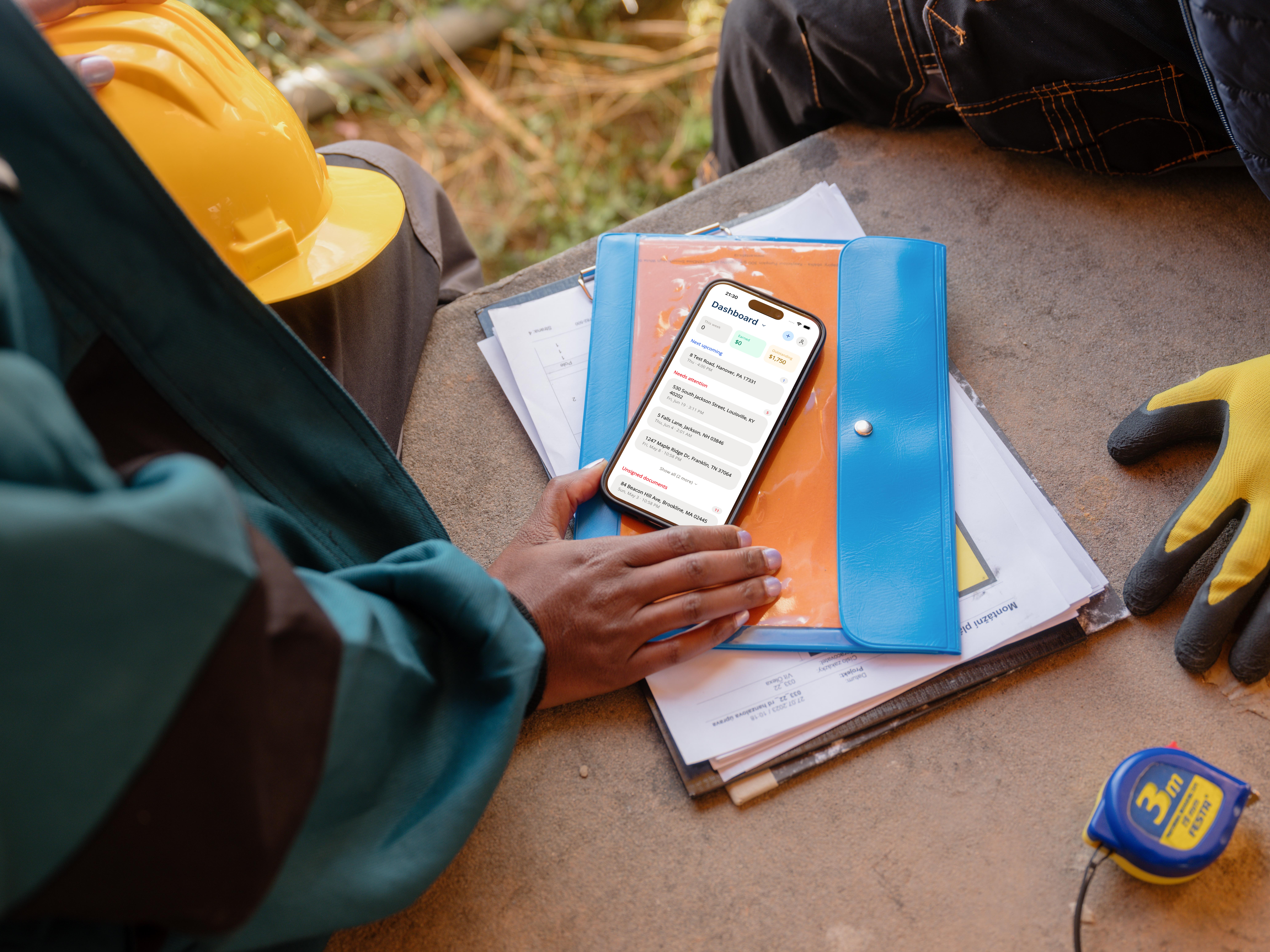 Home inspector at a job site reviewing findings on a phone, hard hat and tape measure beside the paperwork
