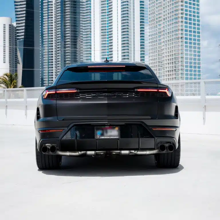 Rear view of a matte black Lamborghini Urus SE, highlighting its distinctive taillights and quad exhaust pipes, with city buildings in the background.
