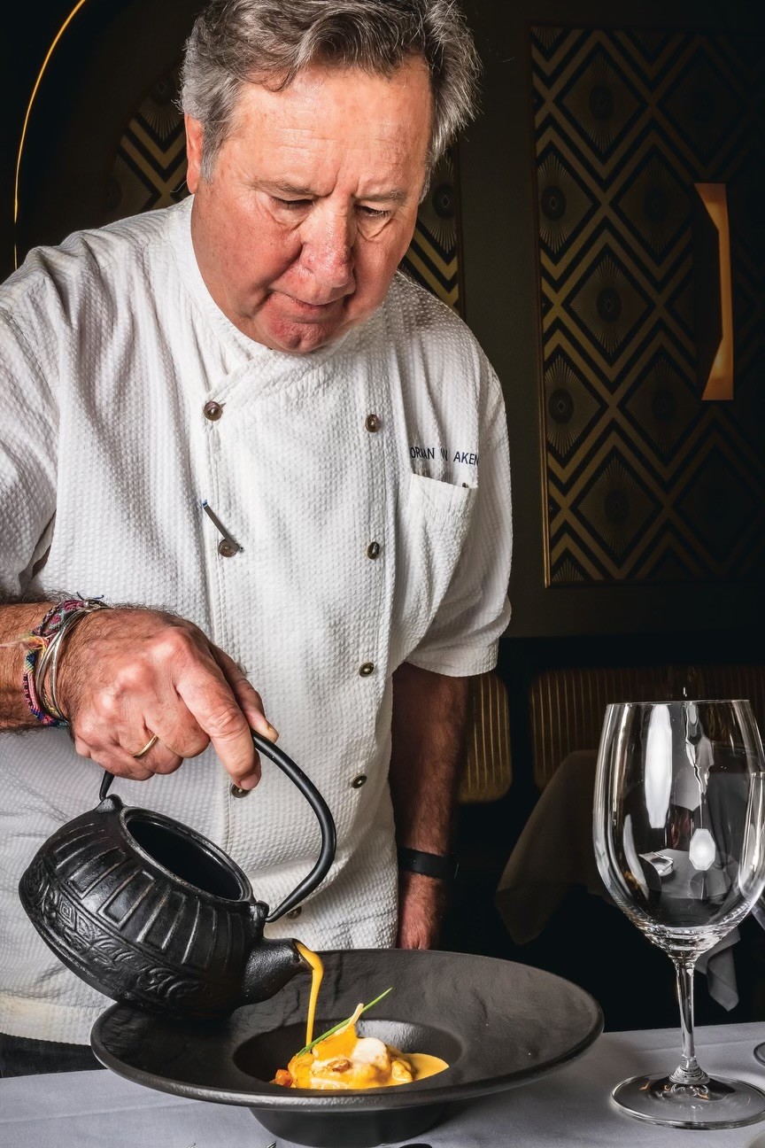 Long, elegant dining table set for a private event. Dark green chairs and yellow banquettes line the room with modern lighting fixtures.