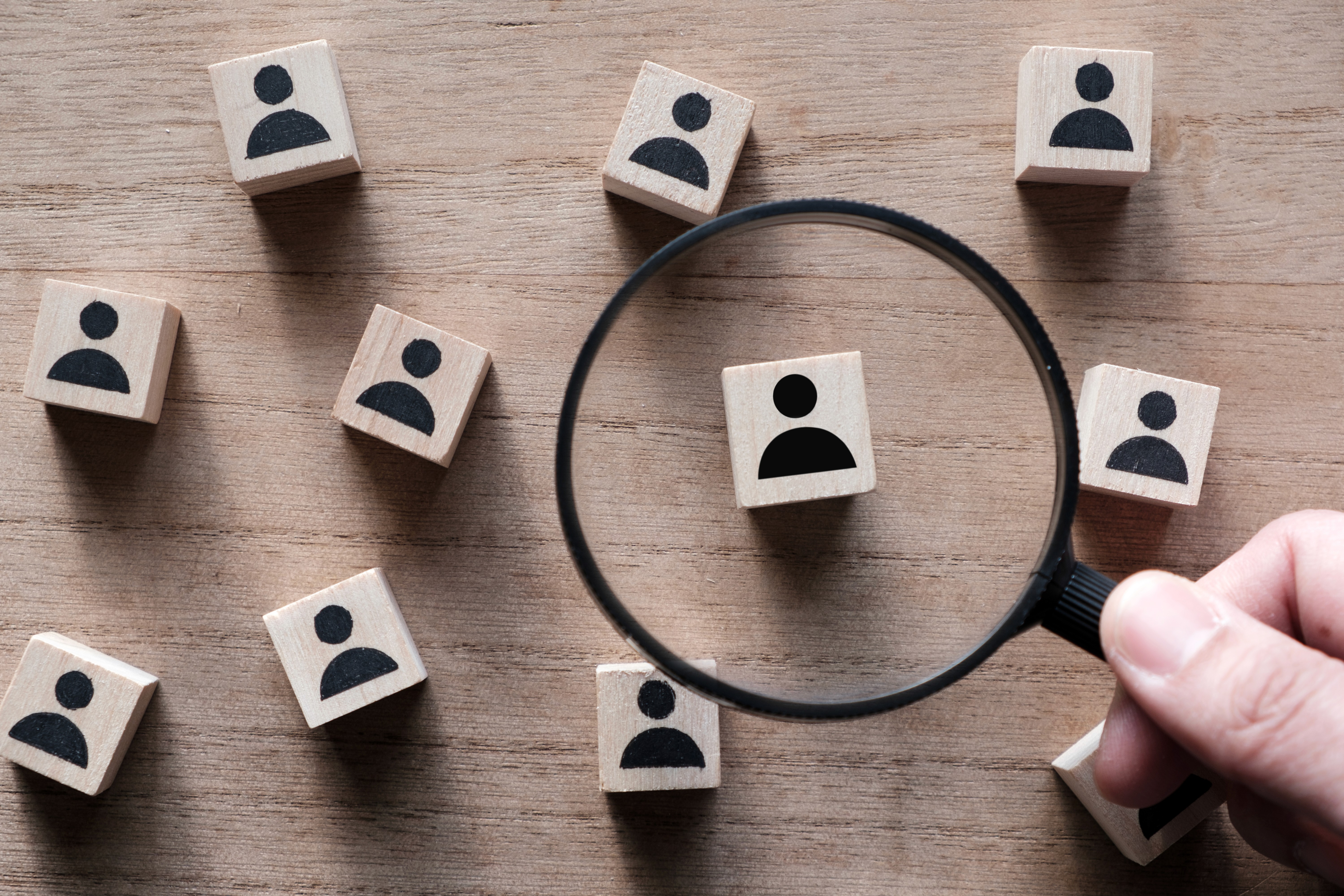 Hand holding a magnifying glass over wooden blocks with person icons on a wooden surface, representing the process of identifying your ideal client and understanding exactly who your content should be speaking to