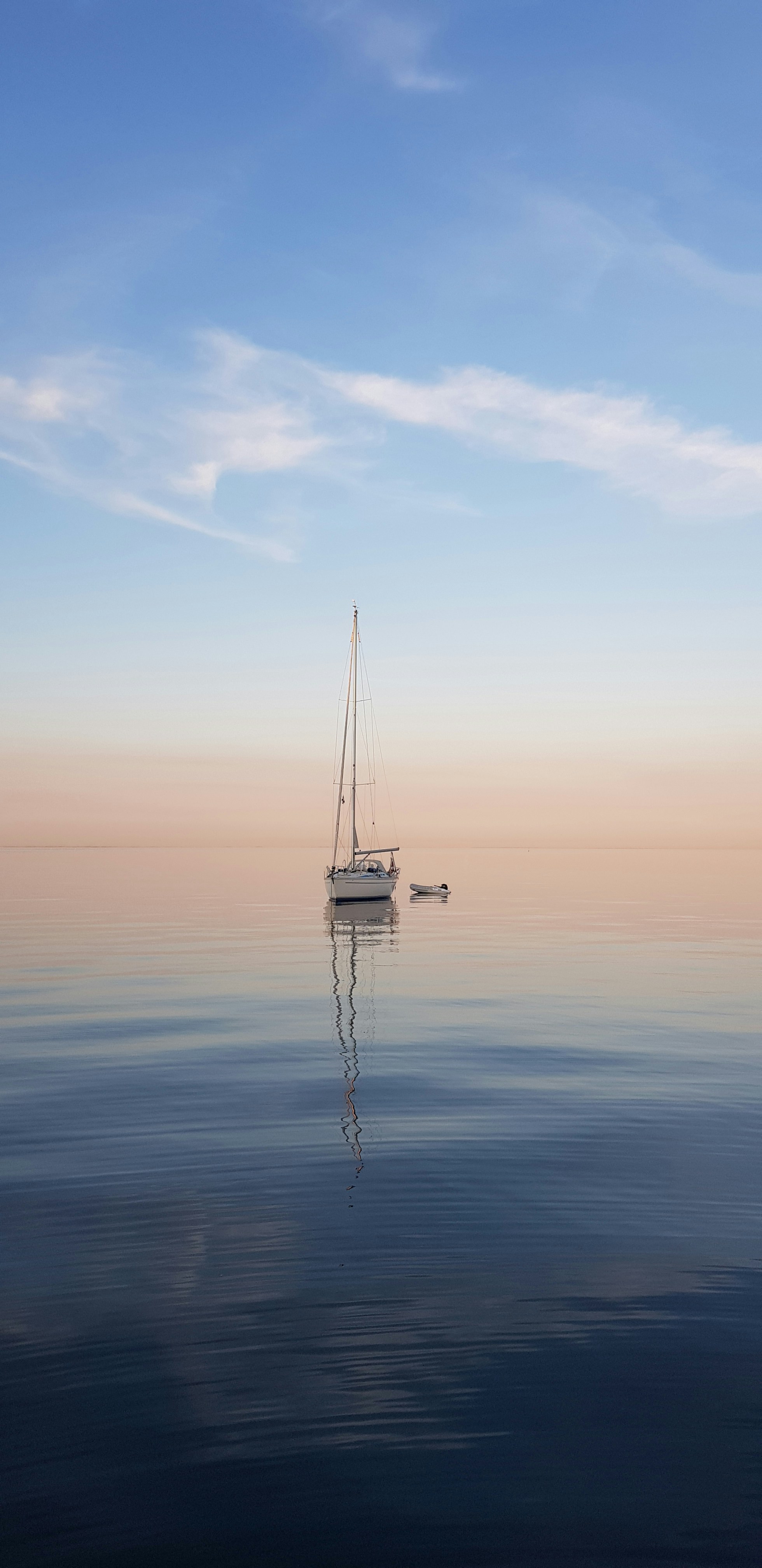 A serene sailboat with its reflection glides on a calm, mirror-like sea under a pastel-colored sky, evoking a peaceful coastal landscape at sunset.