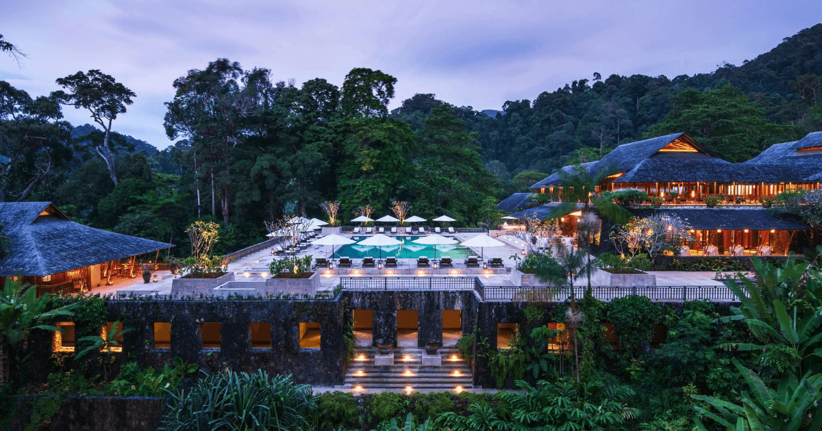 A resort featuring a large terrace with pool, sun loungers, and softly lit buildings surrounded by greenery.