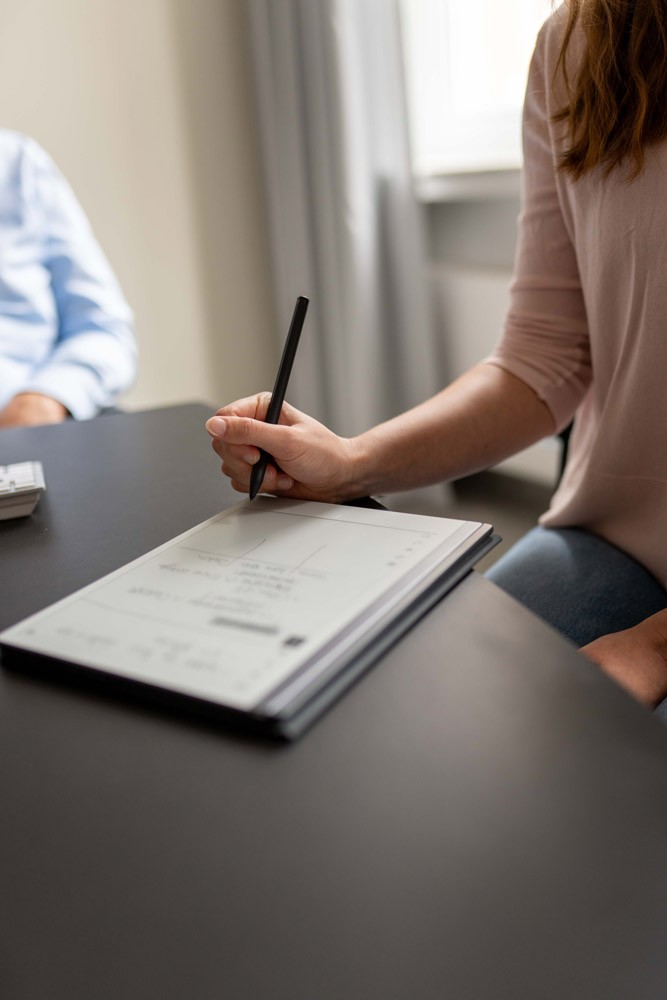 Hand einer Person schreibt mit einem Stift Notizen auf ein Blatt Papier auf einem Schreibtisch.