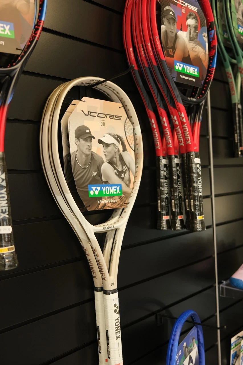Eine Auswahl an Yonex Tennisschlägern, darunter weiße VCORE 100L-Modelle und rote VCORE 100-Schläger, präsentiert an einer schwarzen Wandhalterung. Die Schläger haben ein Werbe-Etikett mit Tennisspielern und dem Yonex-Logo. Im Hintergrund sind weitere Tennisschläger sowie Zubehör wie Overgrips sichtbar.