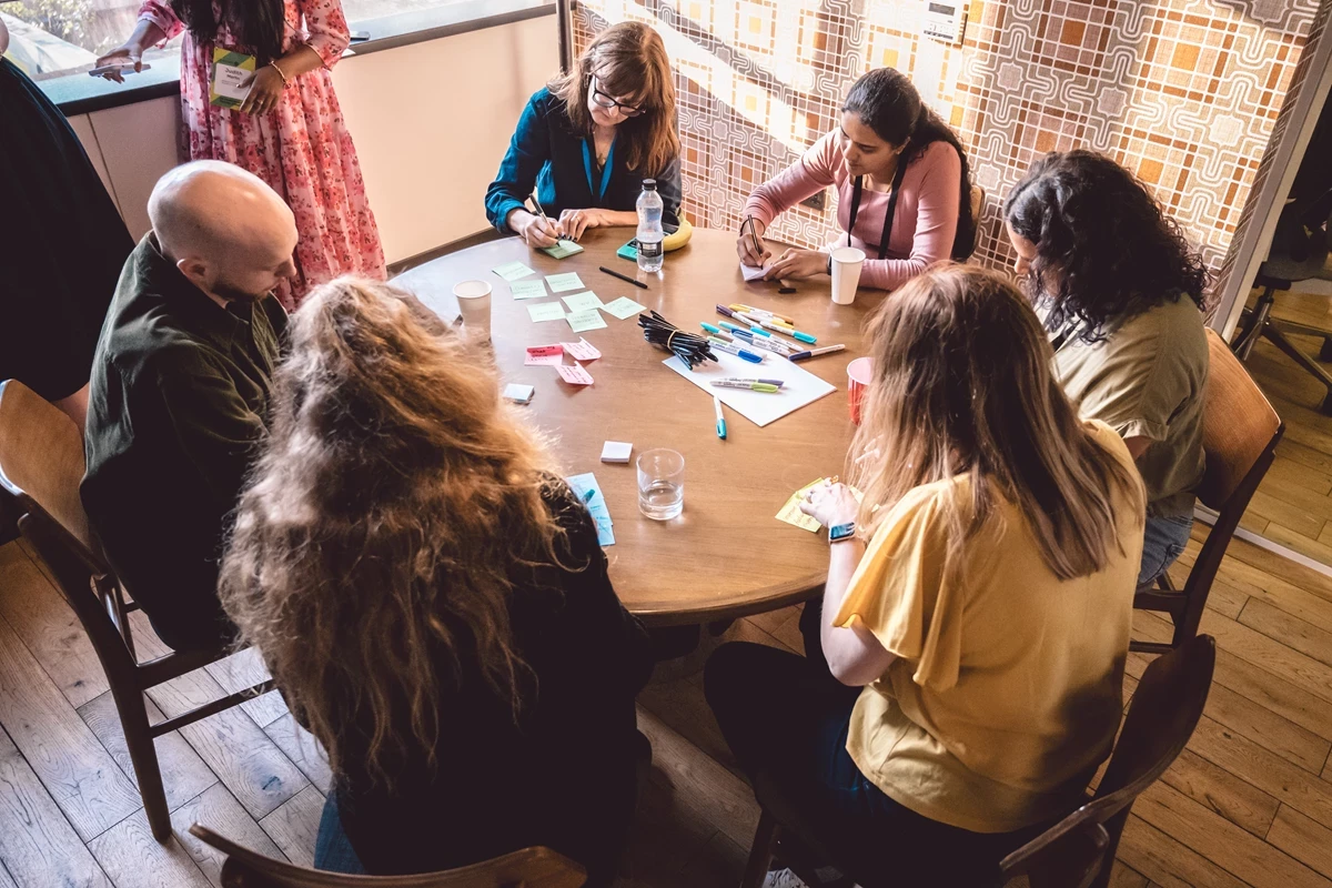 Group of people seated around a wooden table engaged in a collaborative activity with colorful sticky notes, pens, and drinks.
