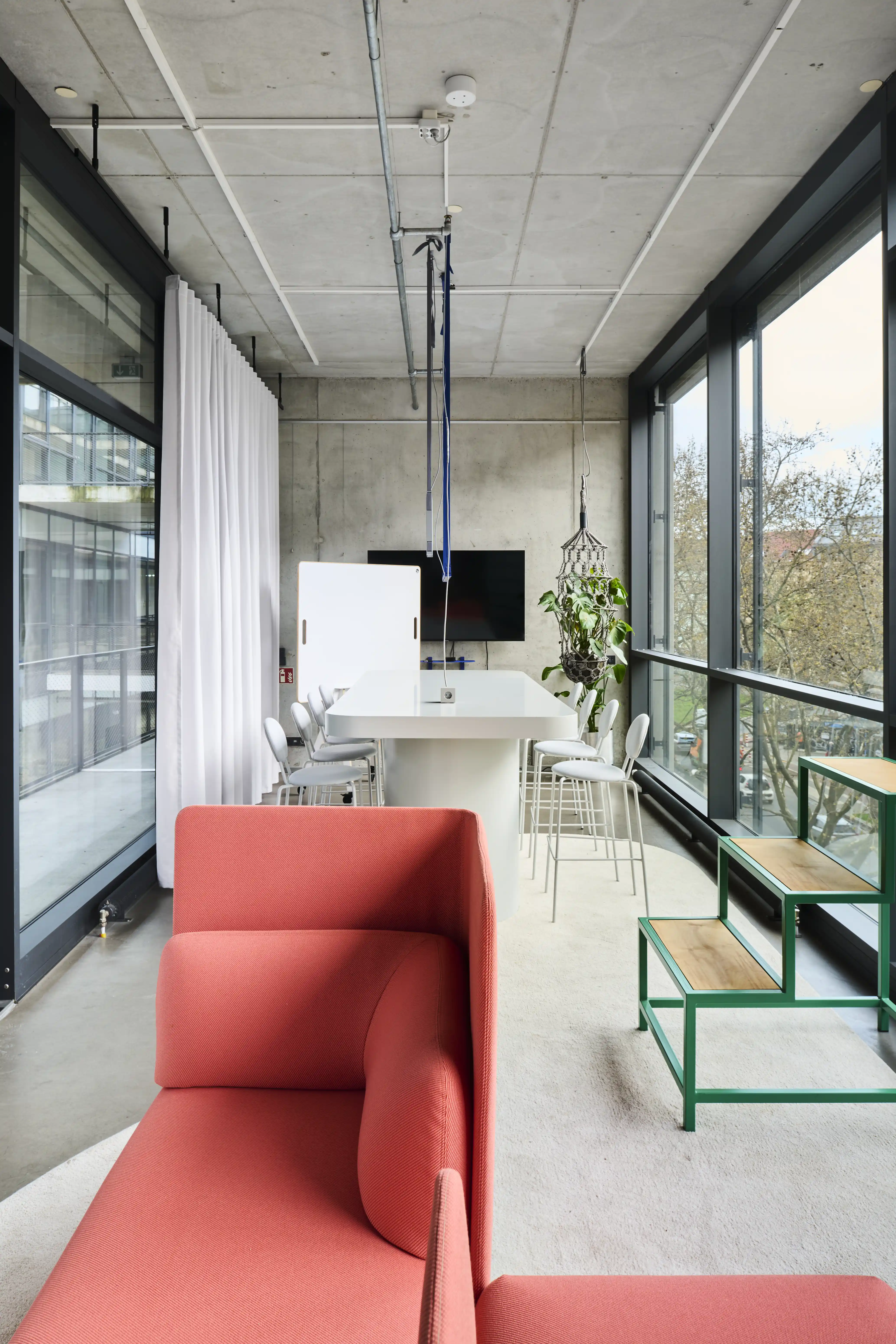 View through the UNDPLUS SHOWROOM with a coral sofa in front, green wooden step stools, white curtains and the high-table meeting zone by the window front.