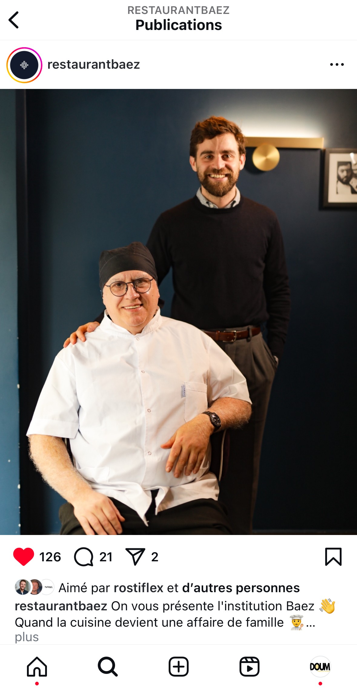 Un post instagram avec deux personnes, l’une assise et l’autre debout, sourient chaleureusement dans une pièce faiblement éclairée aux murs bleu foncé ornés de tableaux encadrés, créant une ambiance cosy et accueillante, empreinte de convivialité.