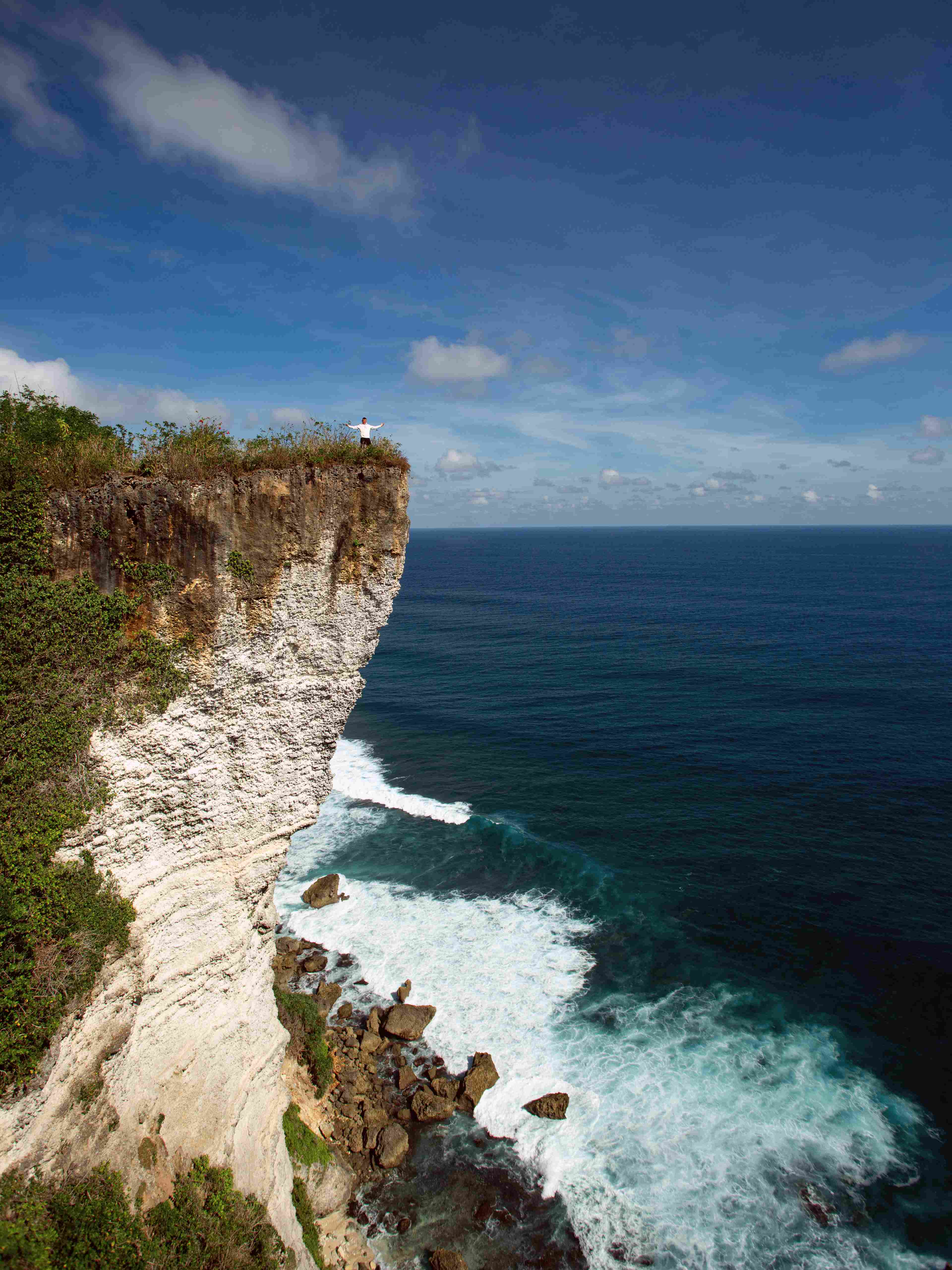 Dramatic ocean view in Uluwatu with a large rock cliff on the left. A person in white stands at the cliff's edge, surrounded by lush greenery and deep blue sea.