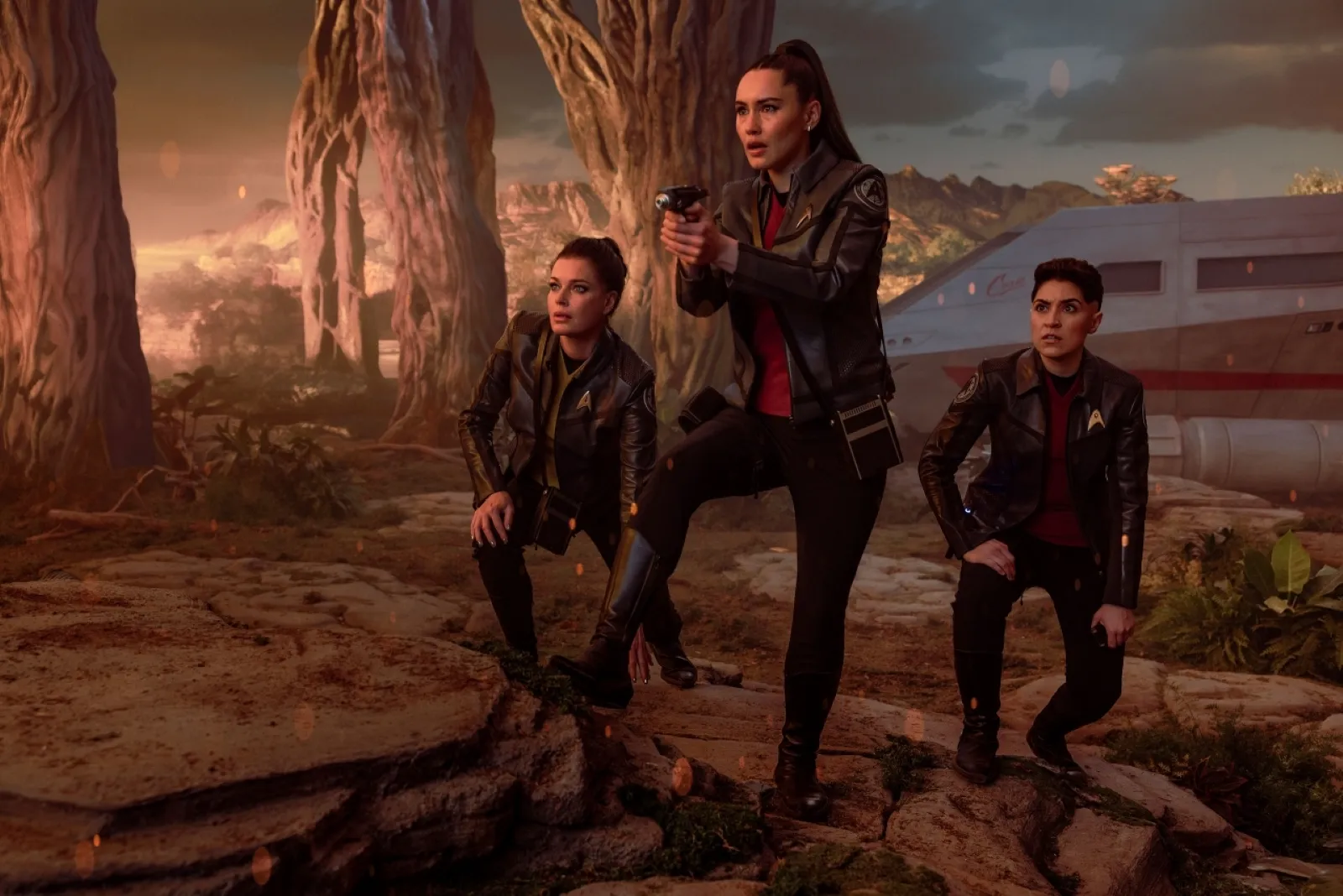 Three Star Trek SNW characters in dark leather attire, resembling a futuristic setting, crouch on rocky terrain under a dramatic sky, with a large, modern vehicle and towering trees in the background.