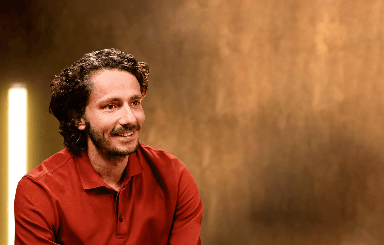 Portrait professionnel et souriant de Guillaume Pley, fondateur du média digital Legend, dans son studio d'enregistrement. Il est vêtu d'un polo rouge terre cuite. Le sujet regarde vers la droite, laissant un large espace négatif à droite de l'image. L'arrière-plan est un mur texturé, flou, dans des tons chauds (or, bronze, marron), éclairé par une colonne lumineuse verticale à l'extrême gauche, créant une ambiance de studio d'interview et de podcast haut de gamme. 