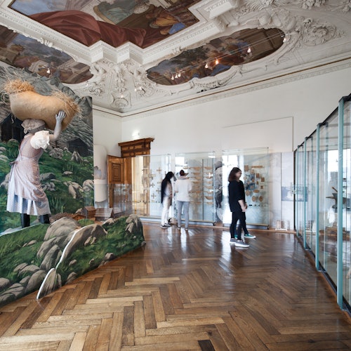 华丽的房间铺有木地板，天花板上有壁画，陈列柜中展示着农场主题的壁画，人们正在参观展览。