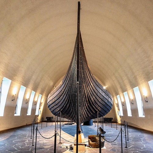 A large, historic Viking ship displayed in a museum with a high arched ceiling and several windows.