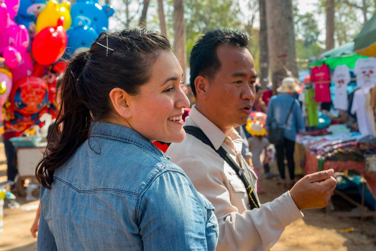 A woman in a denim shirt and a man in a white shirt smile while standing near colorful balloons at an outdoor market.