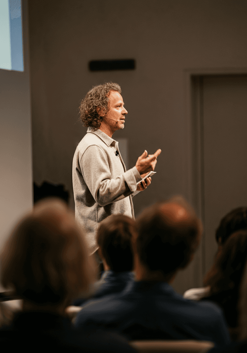 Speaker standing at the front of a room giving a presentation, gesturing with both hands while an audience listens in the foreground.