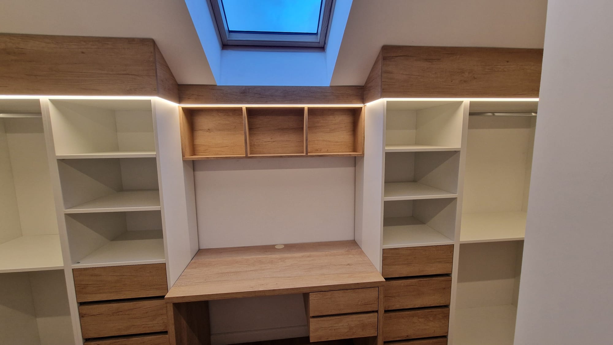 Dressing mansardă cu fereastră Velux, coloane de rafturi albe și sertare din lemn.
