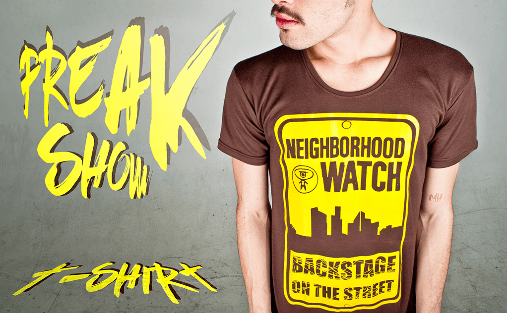Detailed shot of a brown t-shirt featuring a yellow "NEIGHBORHOOD WATCH" sign graphic. Large yellow "FREAK SHOW" typography is placed to the left.