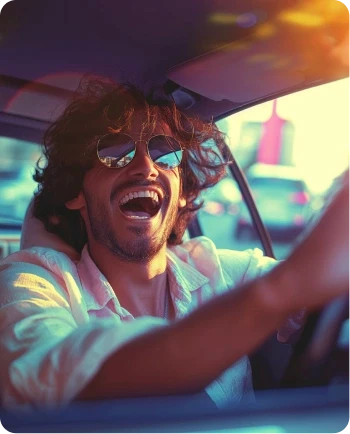 Cheerful young man with curly hair and sunglasses laughing while driving a car during a sunny sunset