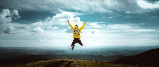 A person wearing a yellow shirt jumps joyfully against a backdrop of blue skies and distant mountains.