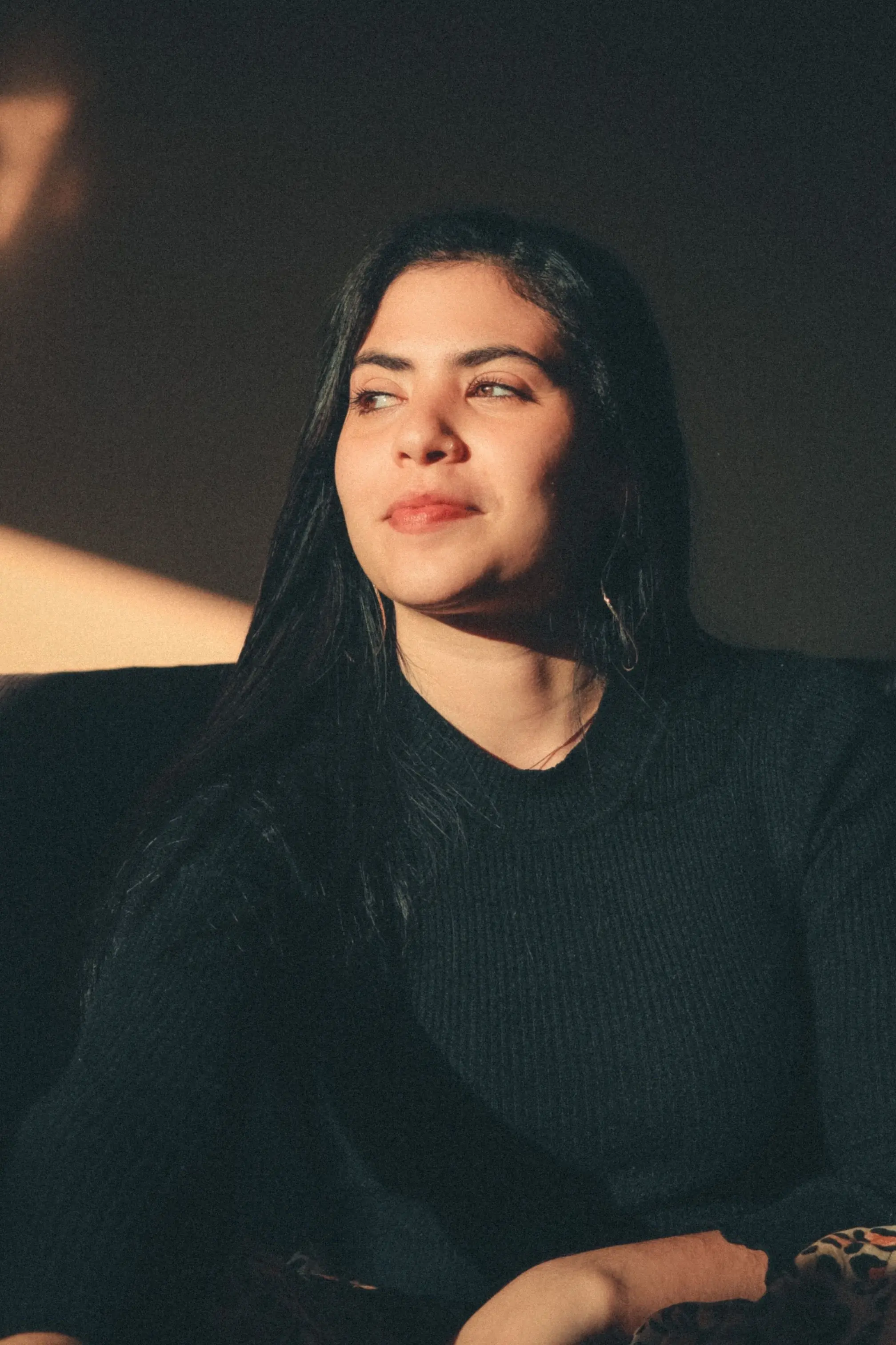 A portrait of a woman with long dark hair wearing a black sweater, illuminated by warm sunlight against a dark background.