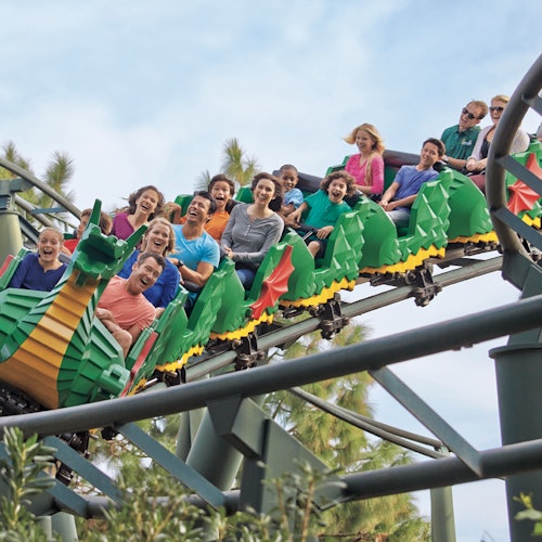 People smiling and raising their hands while riding a colorful dragon-themed rollercoaster with trees visible in the background.