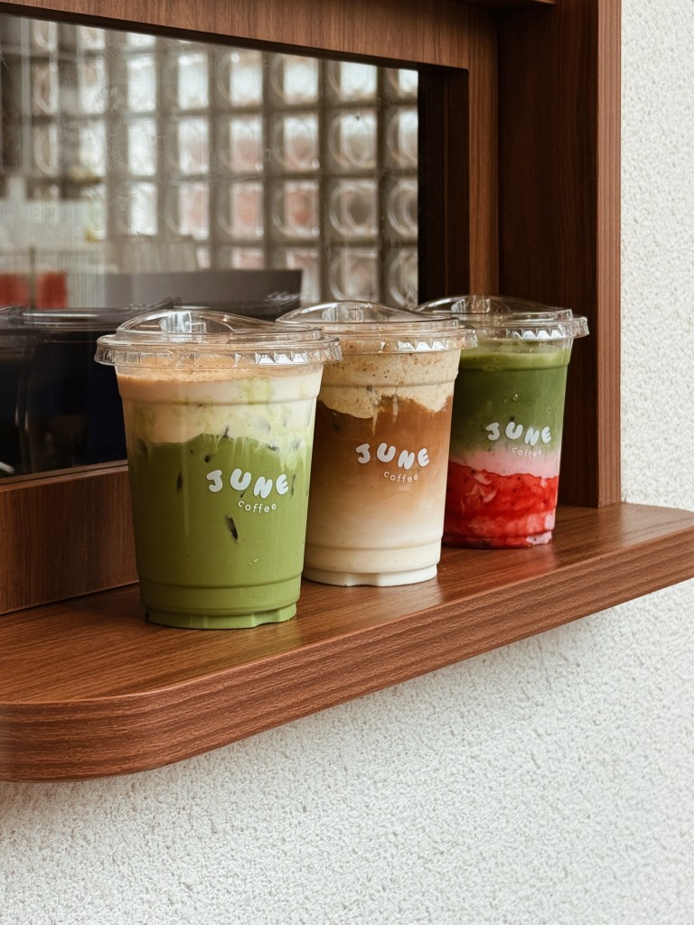 Three cups of iced coffee drinks from June Coffee, displayed on a wooden shelf beside a window.