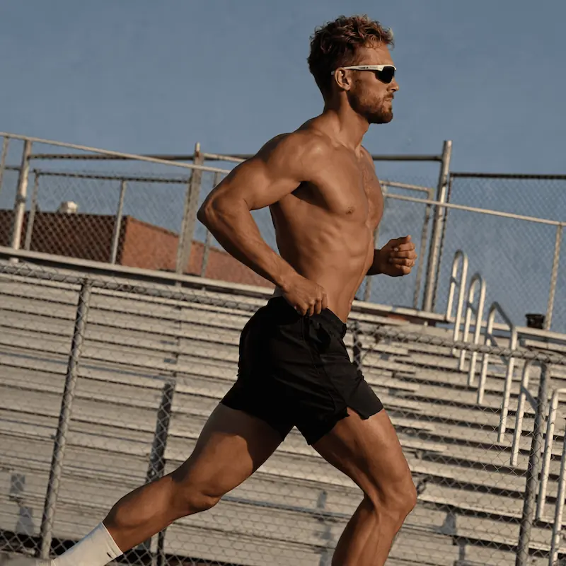 Man running shirtless on a track, wearing black shorts and sunglasses during a workout.