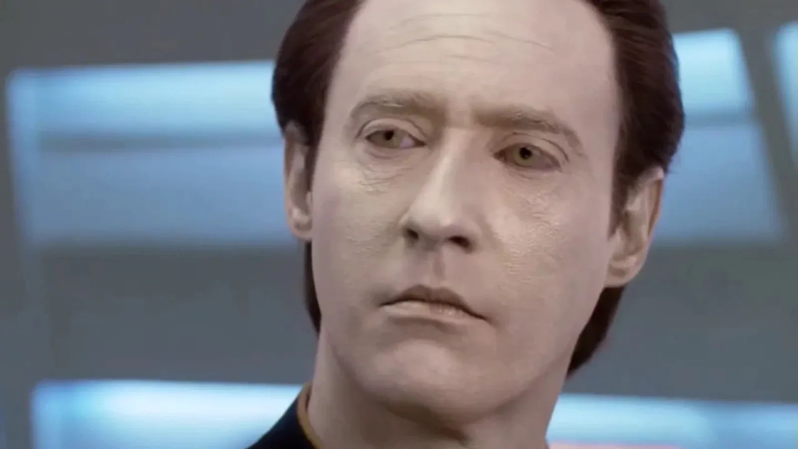 A humanoid character with pale skin and a neutral expression stands against a futuristic, blurred background, likely inside a spaceship or advanced technology setting.