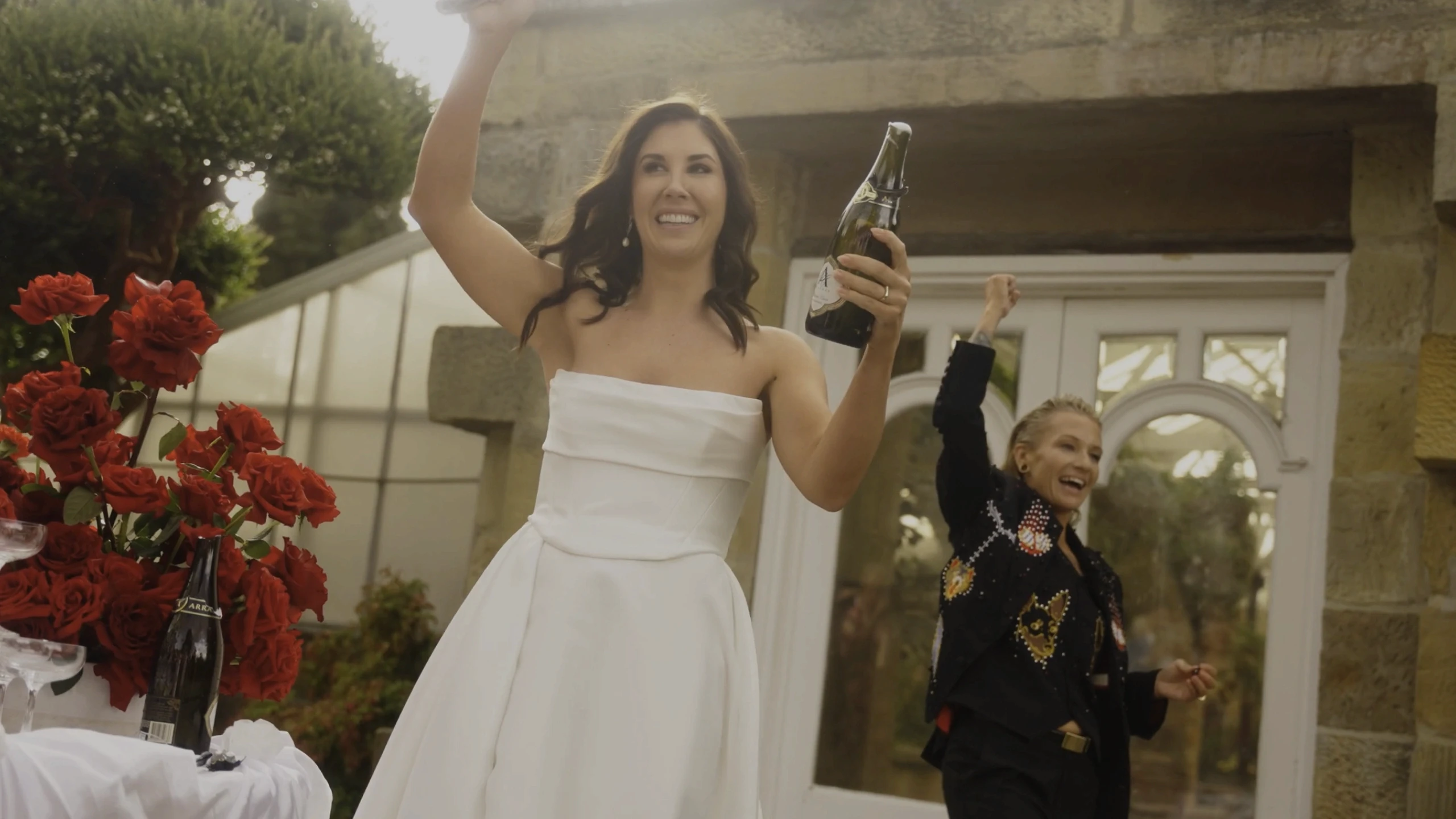 A joyful bride in a strapless white dress holds a bottle while celebrating with friends in a garden setting adorned with red roses and surrounded by a stone building.