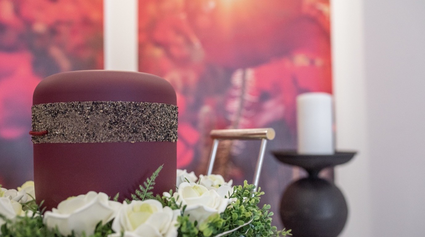 Dunkelrote Urne mit weißem Blumenkranz und Kerze in einem stillen Abschiedsraum