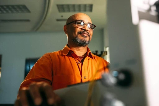 Engineer in an orange shirt and glasses smiling while working at his computer.
