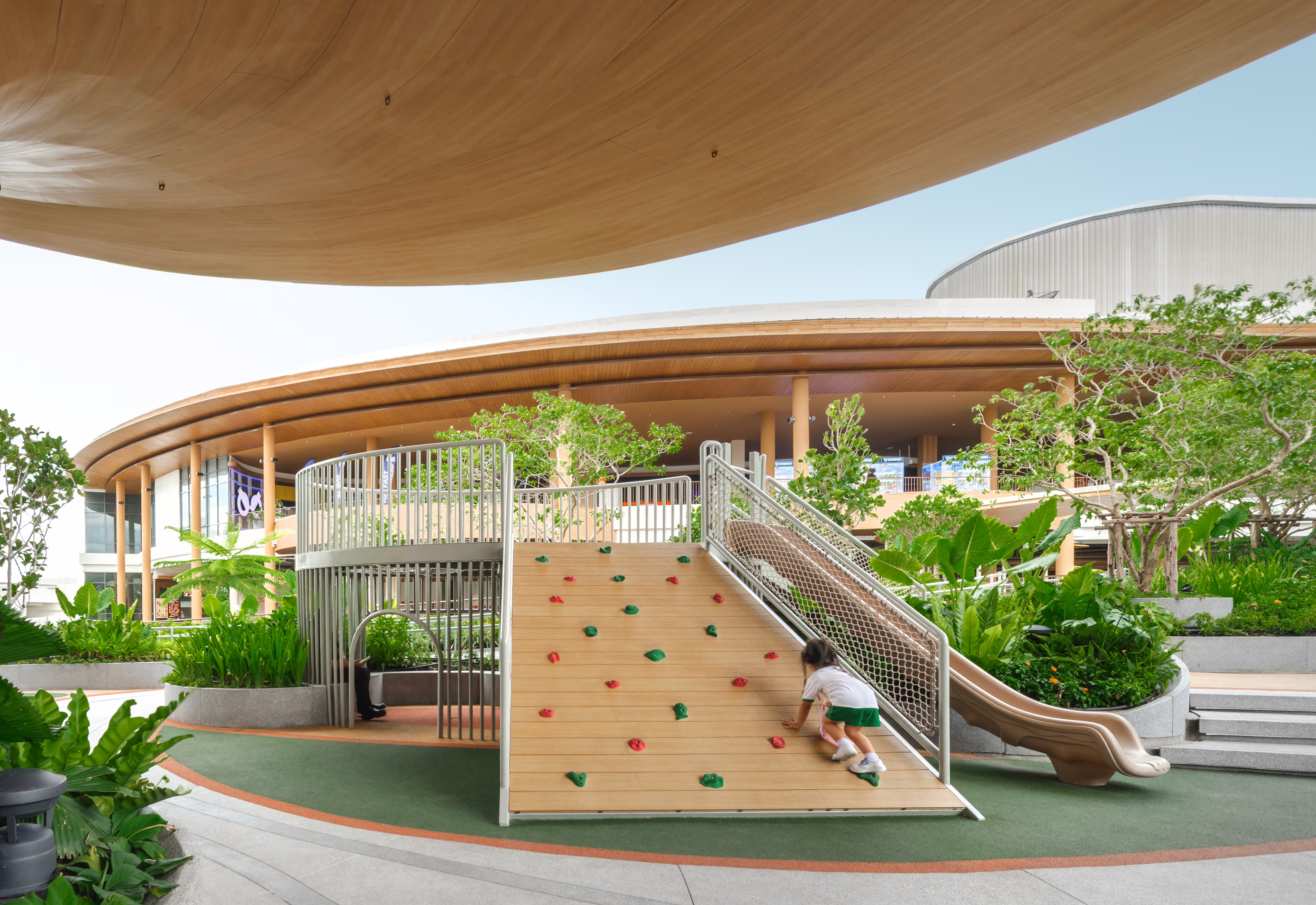 Colorful playground with green structures, a large clock, flowers, and city skyline in the background.
