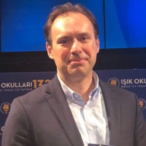 A young person with curly hair smiles at the camera while sitting in front of a laptop against a dark background.