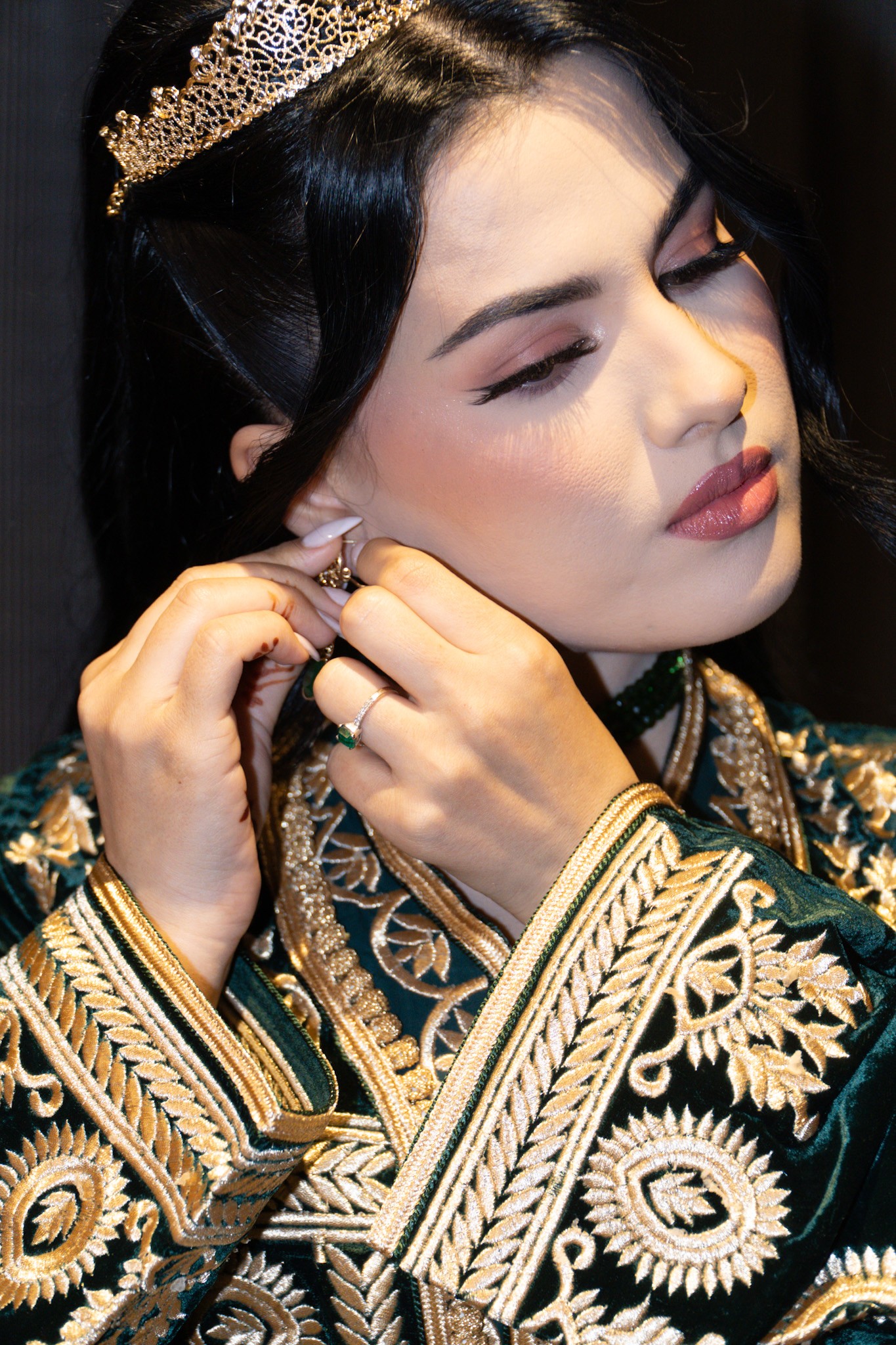 Bride adjusting an earring while wearing a gold headpiece and an embroidered dark robe.