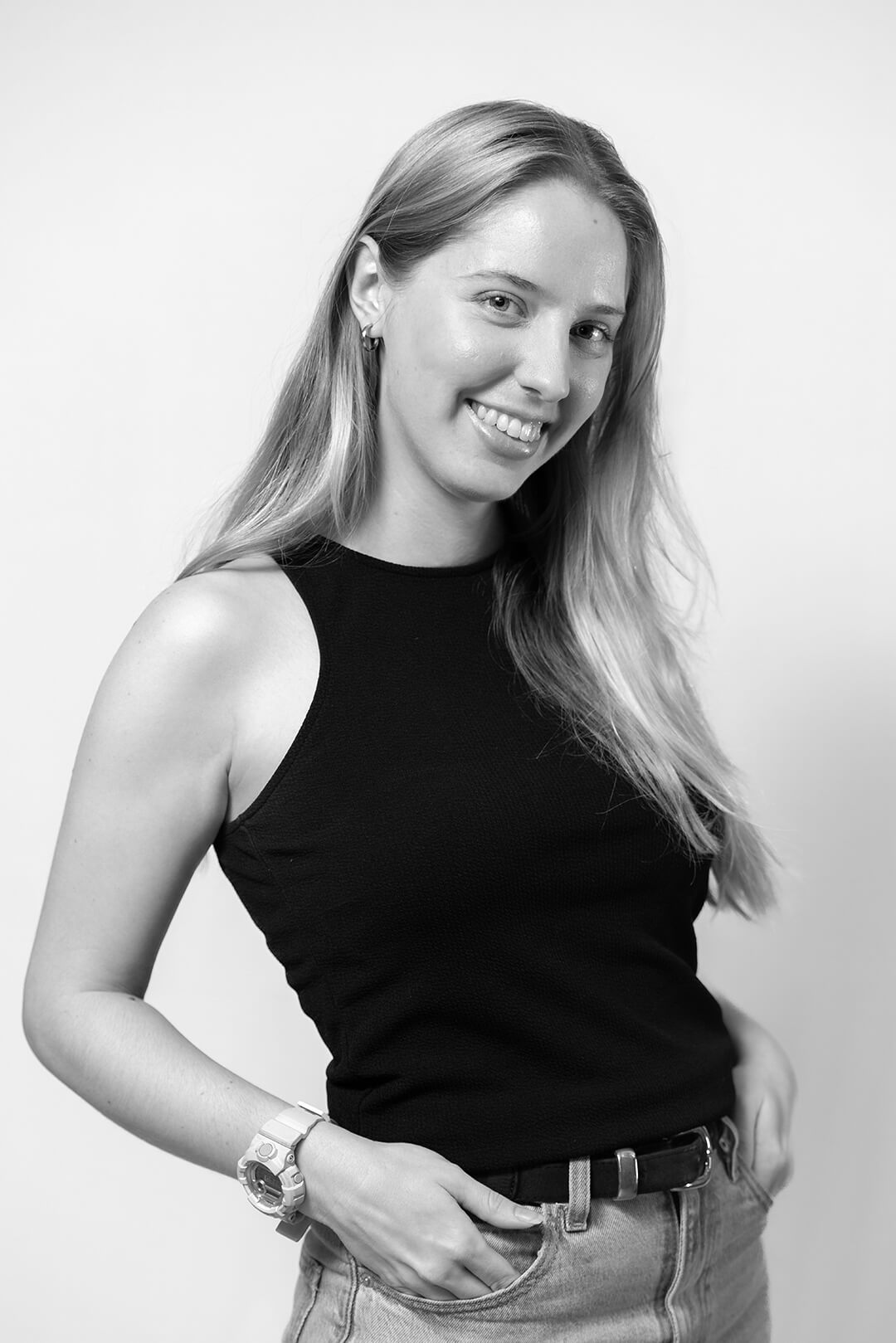 A smiling woman with long hair poses confidently. She's wearing a sleeveless black top, jeans, and a watch. The image is in black and white, conveying a relaxed tone.