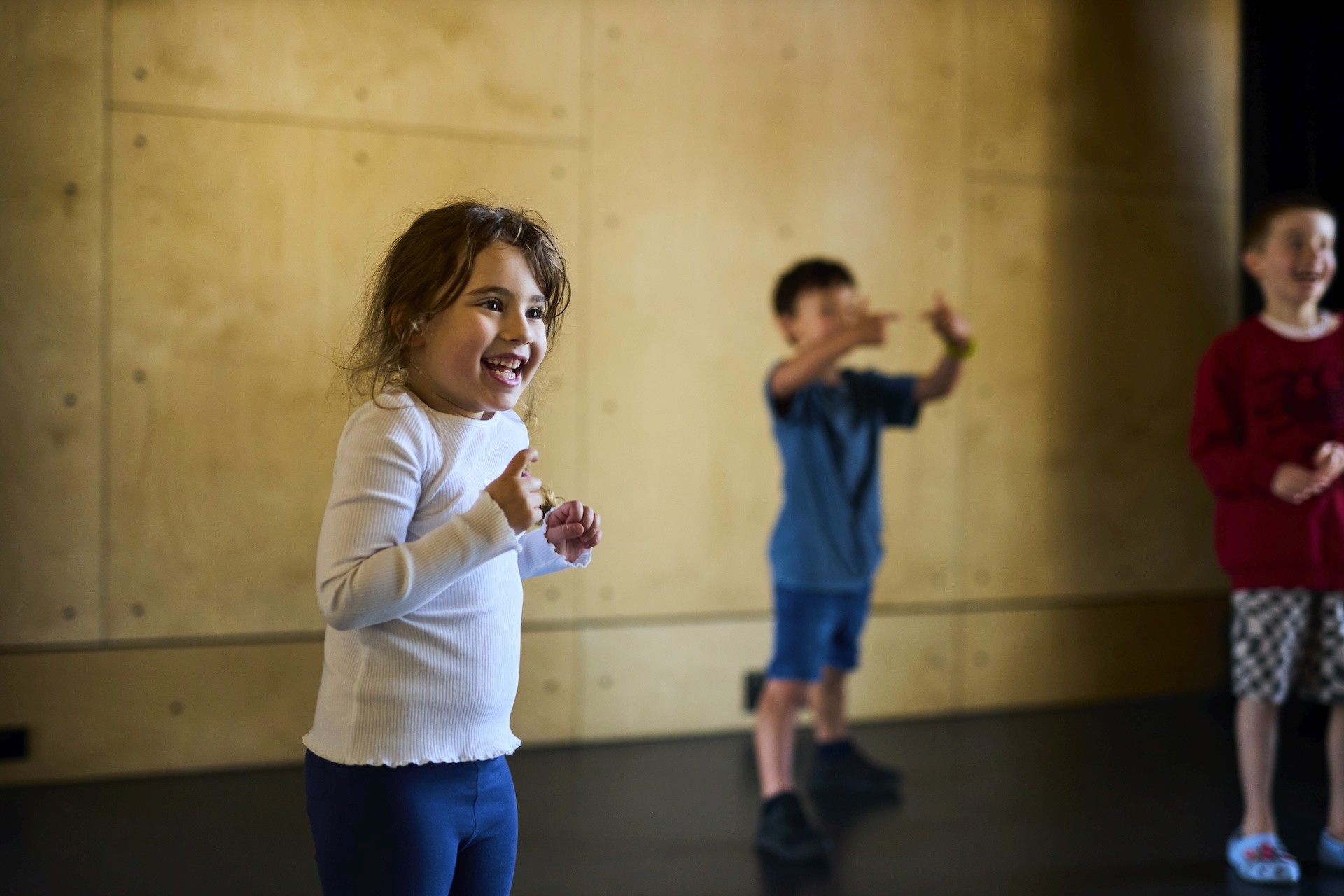 A young child smiles while participating in a movement activity in a studio, with other children blurred in the background copying gestures.