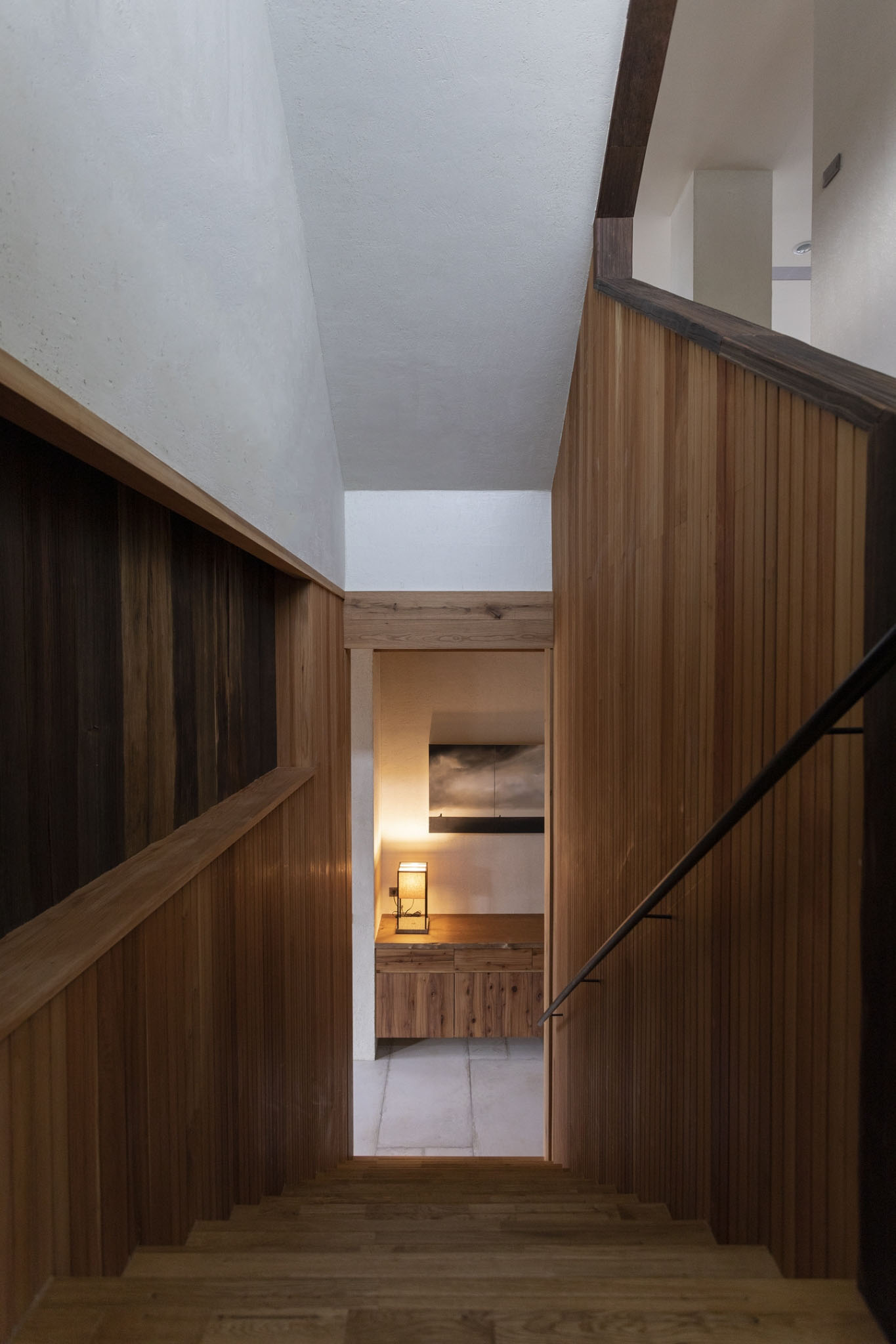 木質牆面與樓梯通往一間擺有橫幅畫作與燈飾的空間，營造出溫潤靜謐的室內過渡感